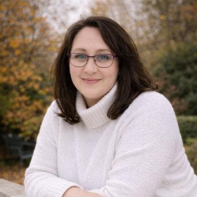 Woman in white sweater smiling at camera outdoors with autumn foliage in background